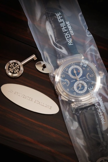 Luxury Patek Philippe watch with protective cover, decorative key, and nameplate on a dark wooden surface.