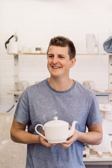 Person wearing a blue shirt smiling while holding a white teapot in a pottery studio background.
