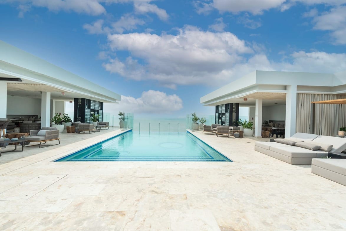 Luxury rooftop infinity pool surrounded by modern lounge seating under a bright blue sky with fluffy clouds