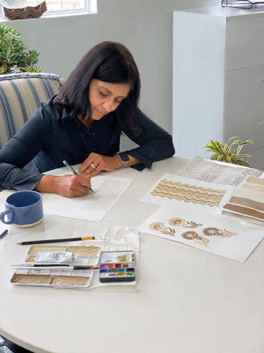 Woman sitting at a table sketching designs on paper, surrounded by art materials and patterns.