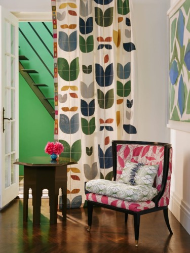 Colorful room with patterned chair, floral pillow, side table with pink flowers, and vibrant leaf-patterned curtain.