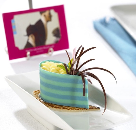 Elegant blue and chocolate-striped dessert on white plate with artistic garnish, beside a colorful illustrated card.