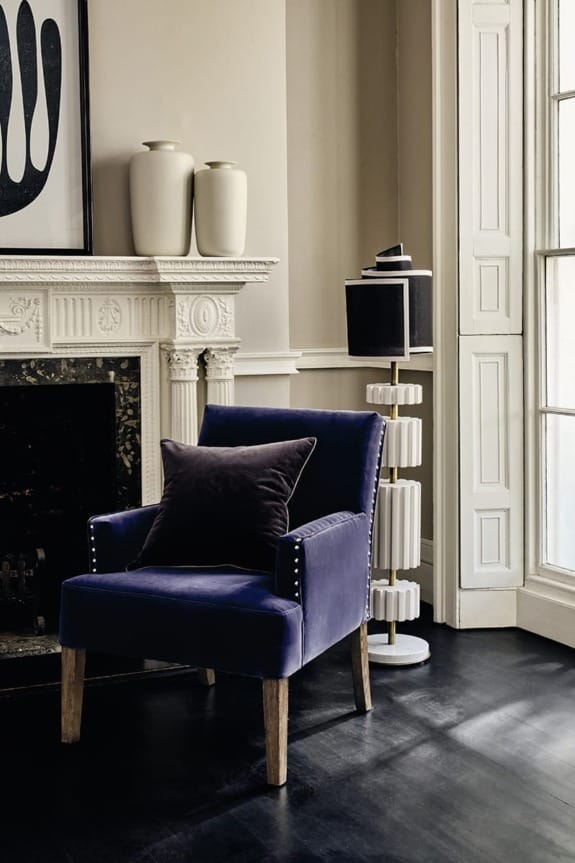 Blue velvet chair with a dark cushion, next to a fireplace and a modern floor lamp in a stylish room.