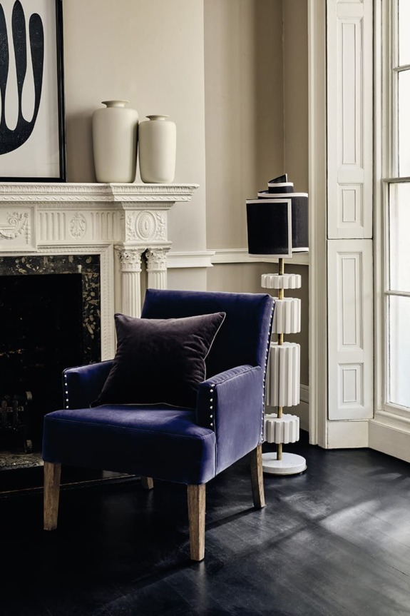 Blue velvet chair with a dark cushion, next to a fireplace and a modern floor lamp in a stylish room.