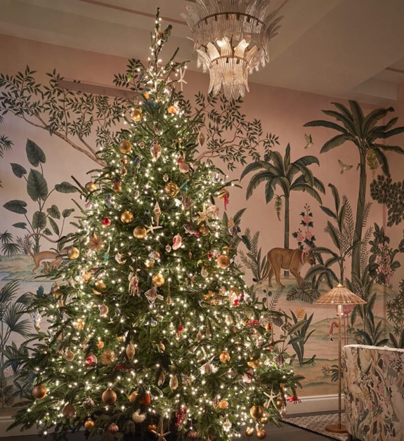 Decorated Christmas tree with lights and ornaments in a room with tropical-themed wallpaper and a chandelier above.