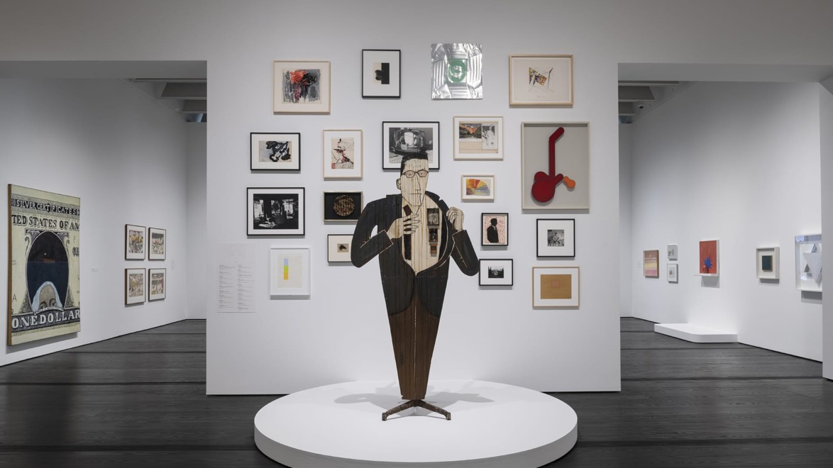 Art exhibit with a saxophonist sculpture in front of framed artworks on white walls in a modern gallery space.