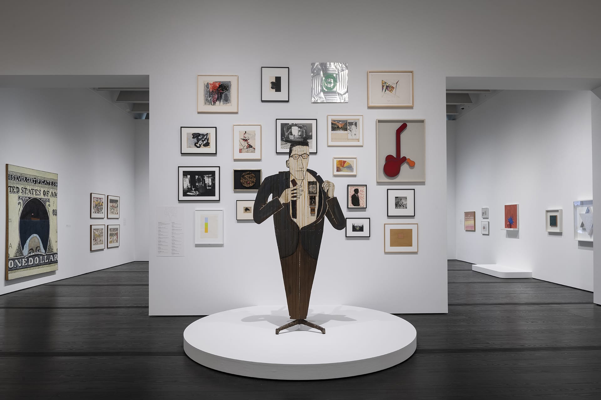 Art exhibit with a saxophonist sculpture in front of framed artworks on white walls in a modern gallery space.