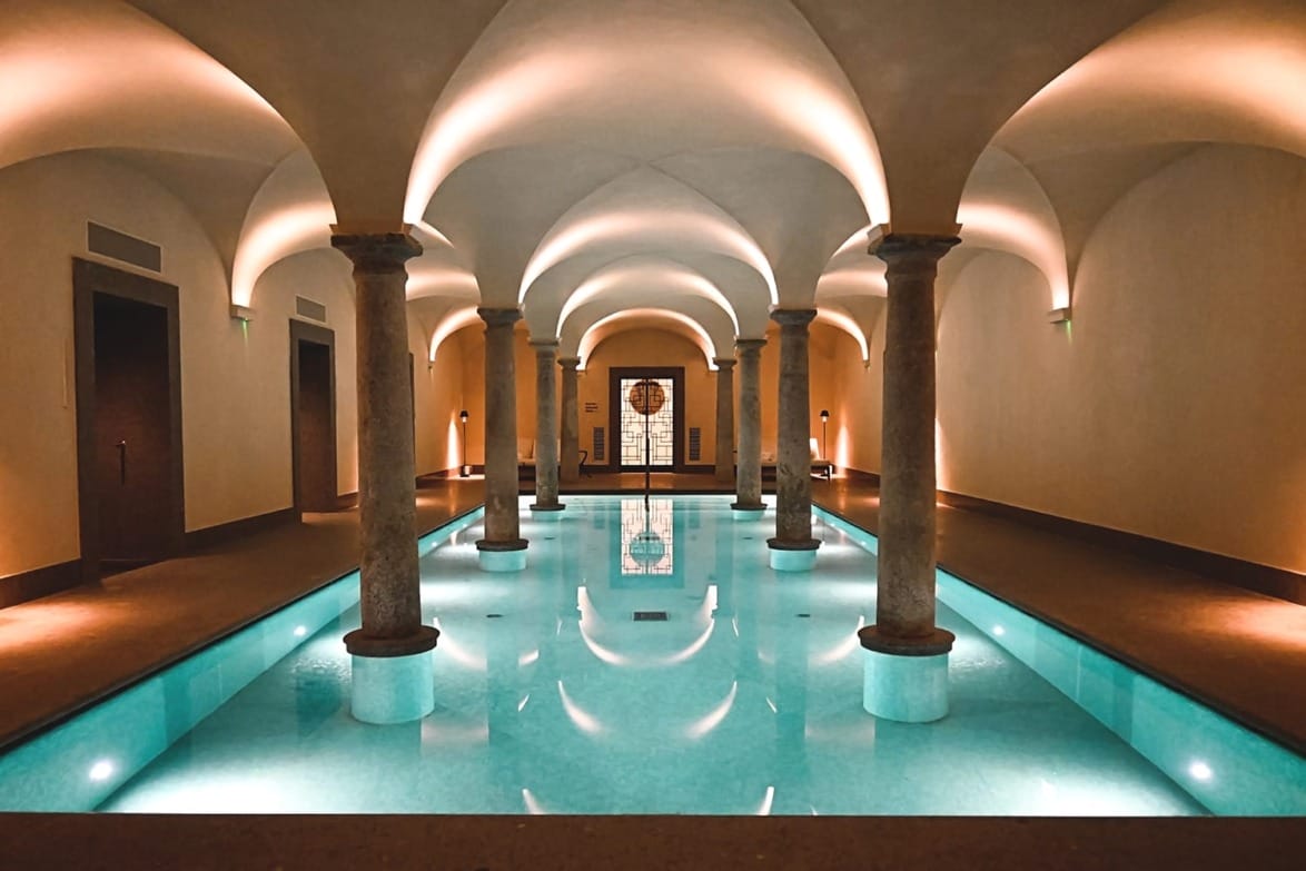 Elegant indoor pool with arched ceilings and illuminated pillars reflecting on the water's surface in luxurious setting.