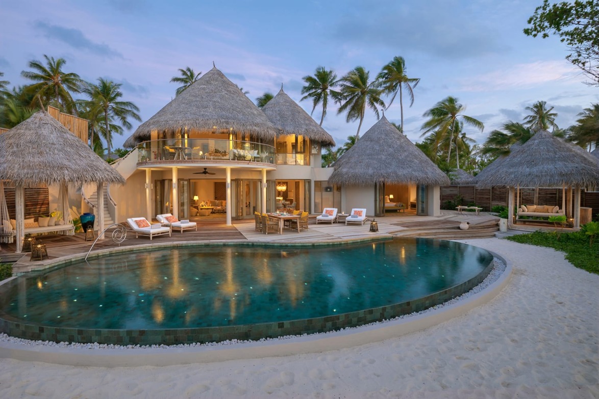 Luxurious beachfront villa with thatched roofs, surrounded by palm trees and a sparkling pool at sunset.