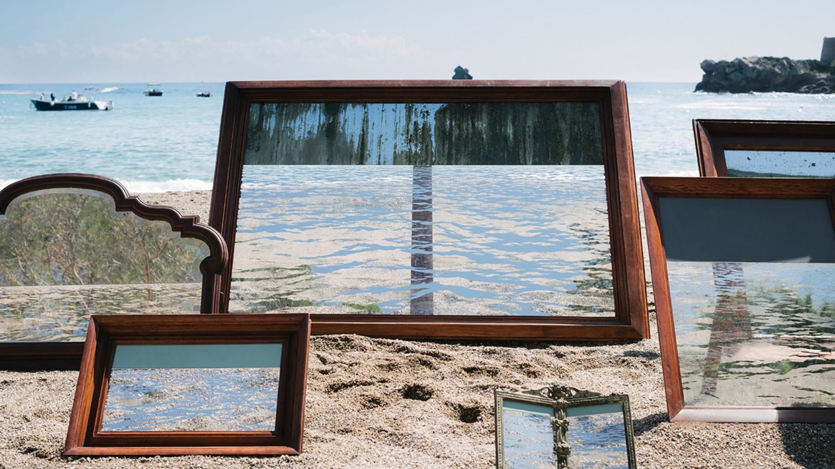 Frames reflecting the ocean and sky placed on a sandy beach with boats in the background.