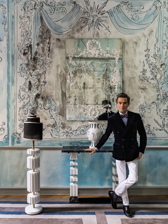 Man in formal attire standing in an elegantly decorated room with ornate wall designs and stylish furnishings.