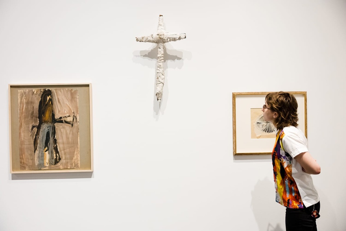 Person observing abstract art pieces in a gallery, including a cross sculpture, wearing a colorful vest.