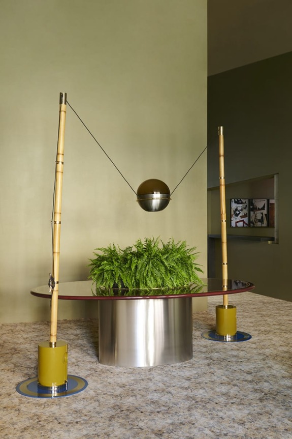 Modern interior with unique light fixture, bamboo poles, and green plants on a sleek metal table against a beige wall.