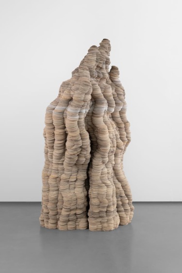 Abstract sculpture made of stacked, textured beige and gray discs, resembling a tall, organic formation in a gallery setting.