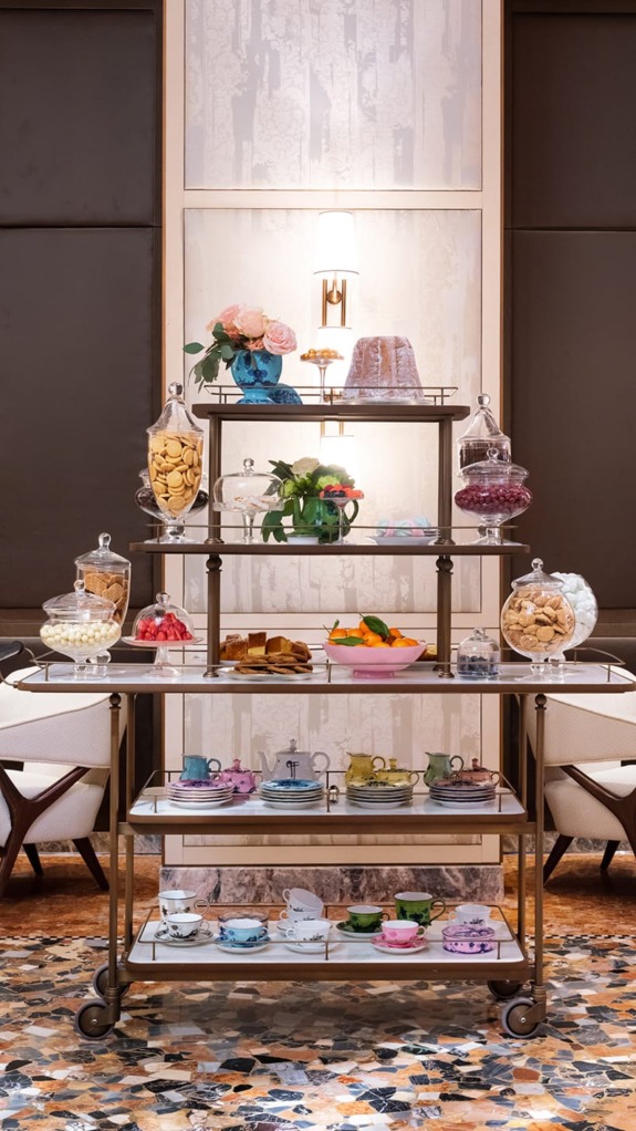 Dessert display on tiered cart with cookies, cakes, flowers, and colorful teacups in elegant room setting.