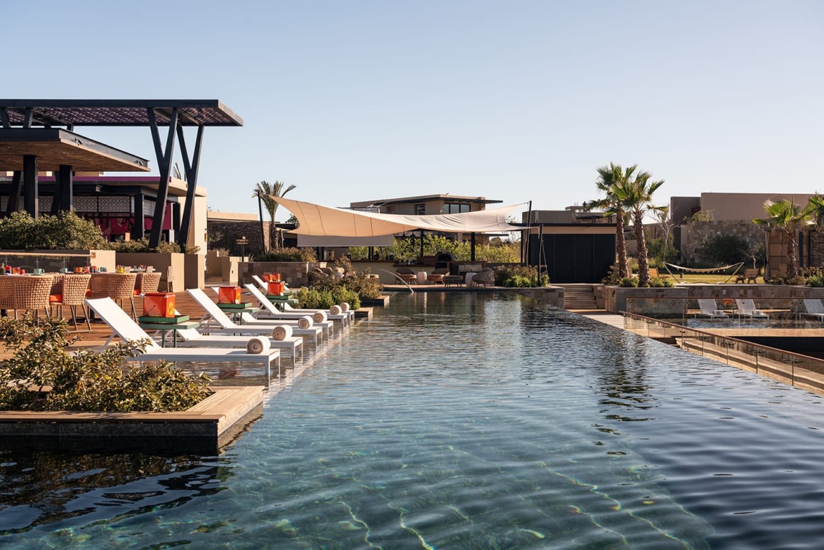 Luxurious resort pool area with lounge chairs, tropical plants, and modern architecture under a clear blue sky.