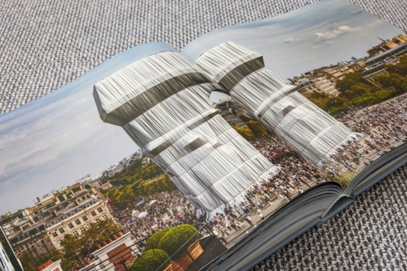 Open book on a gray carpet showing a photograph of large, wrapped towers surrounded by crowds in an urban setting.