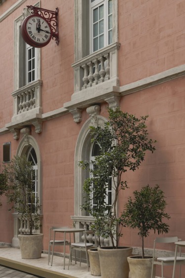 Pink building facade with vintage clock, arched windows, and potted trees on patio.