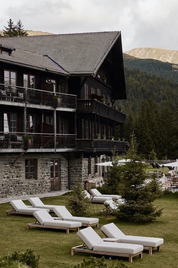 Rustic mountain lodge with lounge chairs on the lawn surrounded by evergreens and hills in the background.