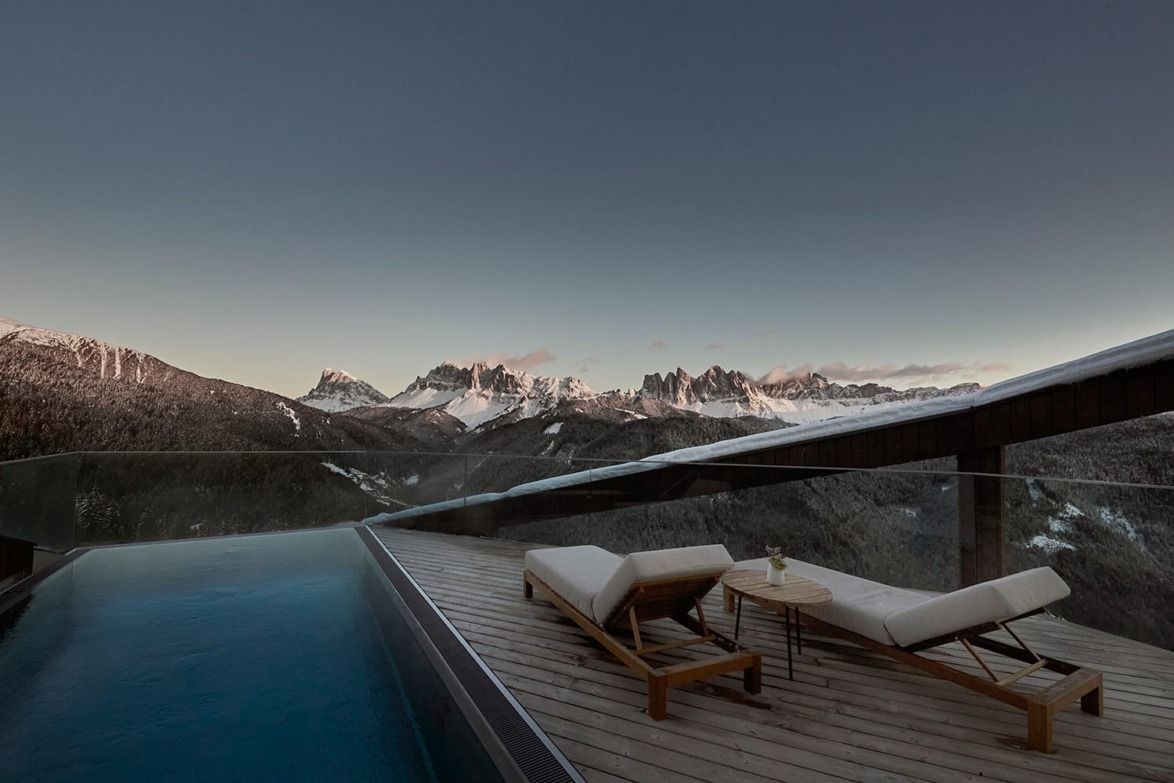 Mountain view from a deck with an infinity pool and two lounge chairs in a tranquil, snowy landscape at dusk