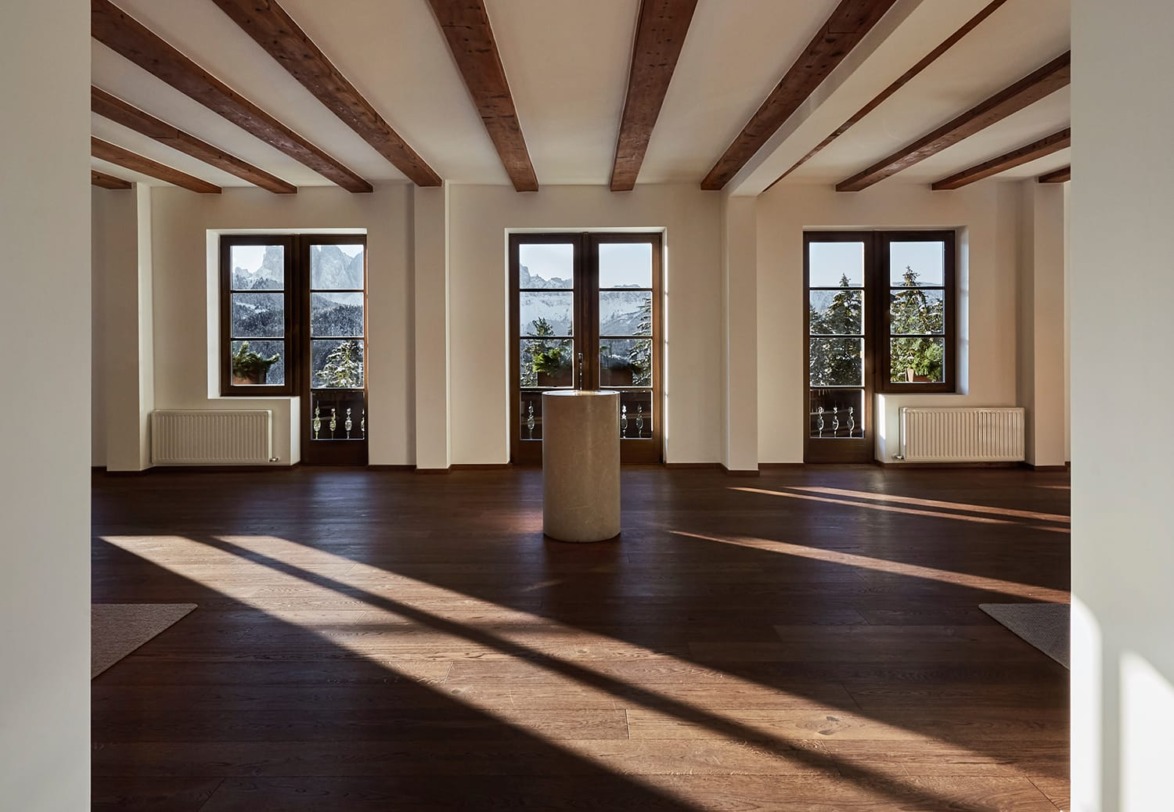Spacious room with wooden beams, large windows overlooking trees and mountains, and sunlight casting shadows on the floor.