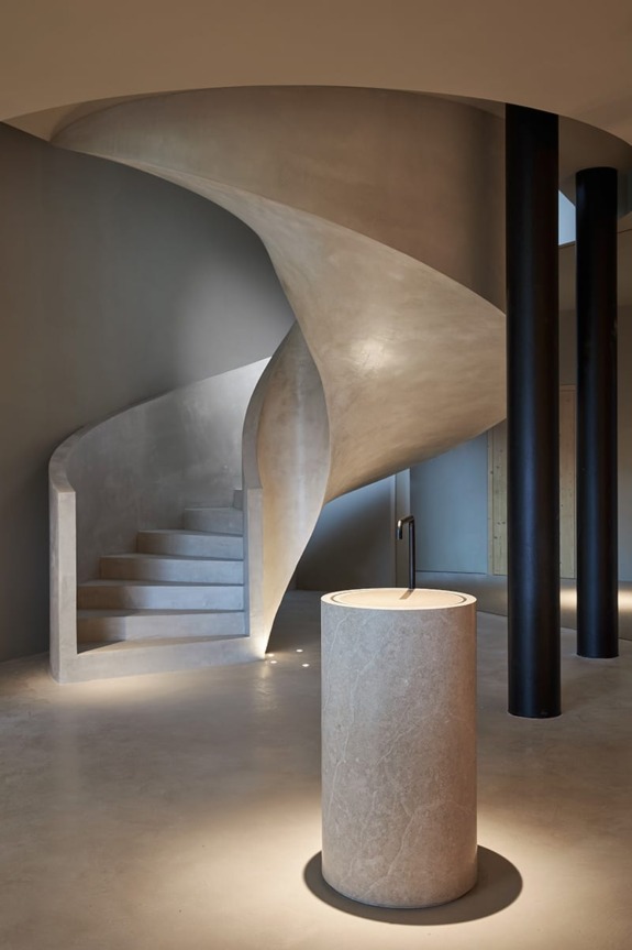 Modern spiral staircase with smooth concrete finish and a cylindrical stone pedestal in a dimly lit interior.