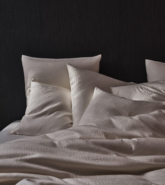 Beige bedding set with textured pillows and blanket against a dark wall background.