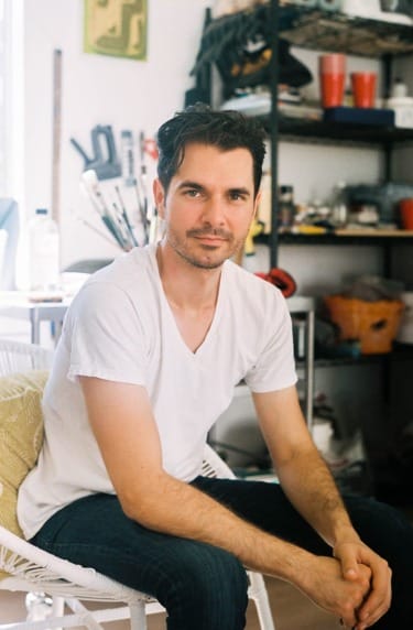 Man sitting in a casual art studio, wearing a white t-shirt and jeans, surrounded by various art supplies and tools.