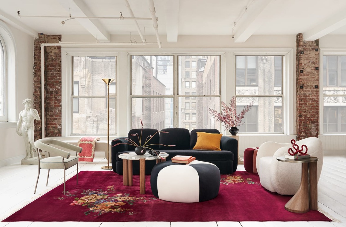 Modern loft living room with eclectic furniture, red floral rug, large windows, and exposed brick walls.