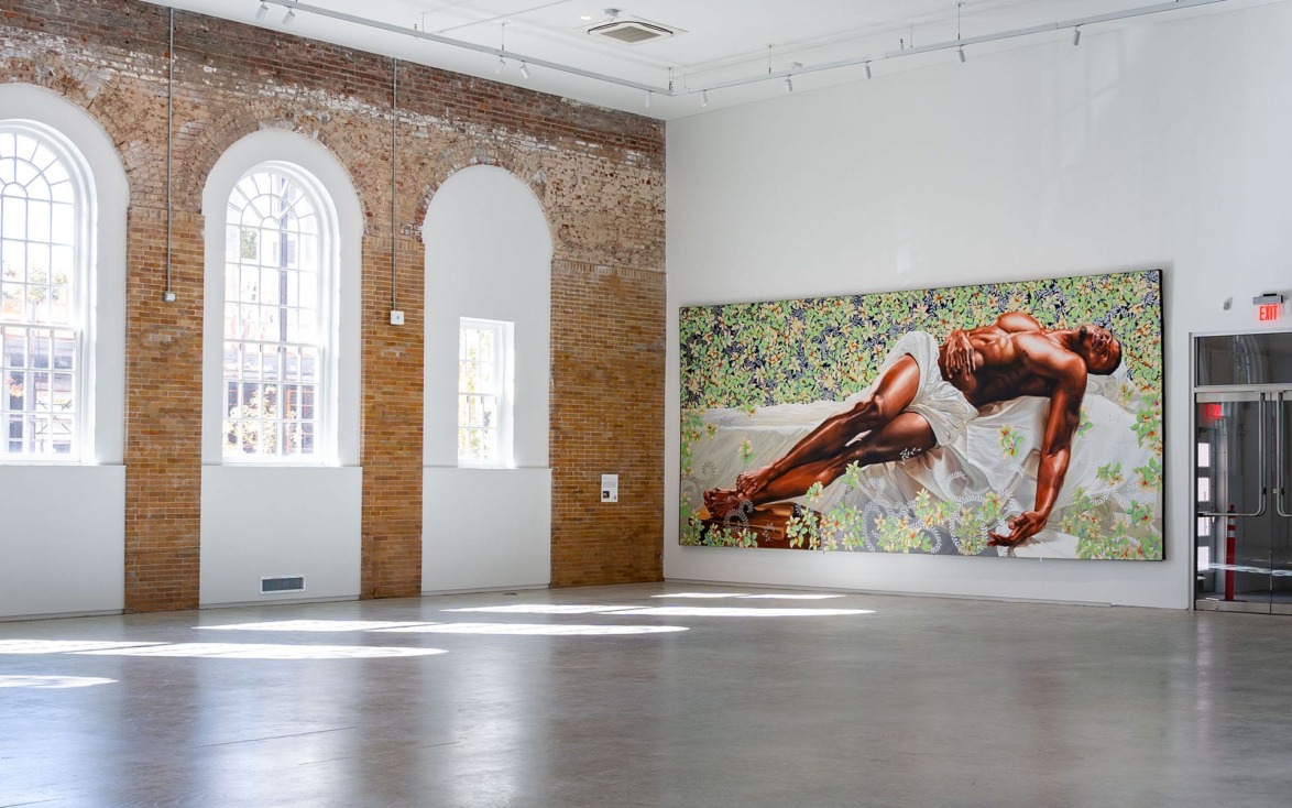Spacious art gallery with brick walls displaying a large painting of a reclining person surrounded by floral patterns.