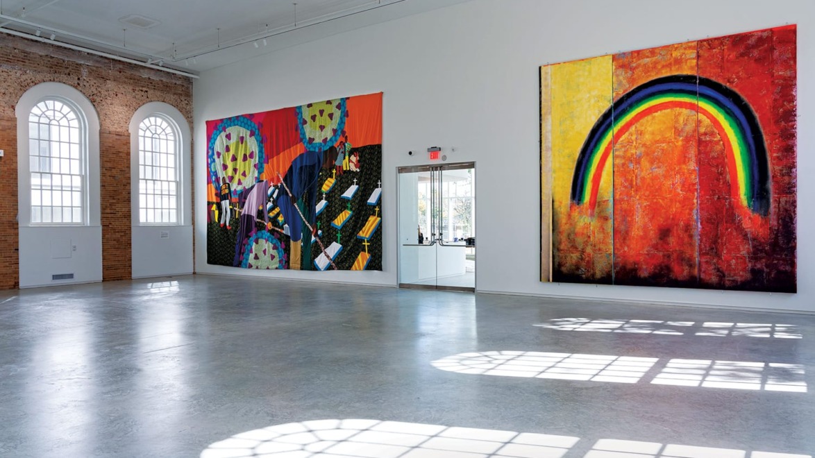 Spacious art gallery with large colorful abstract paintings on white walls and arched windows casting patterned shadows on the floor.