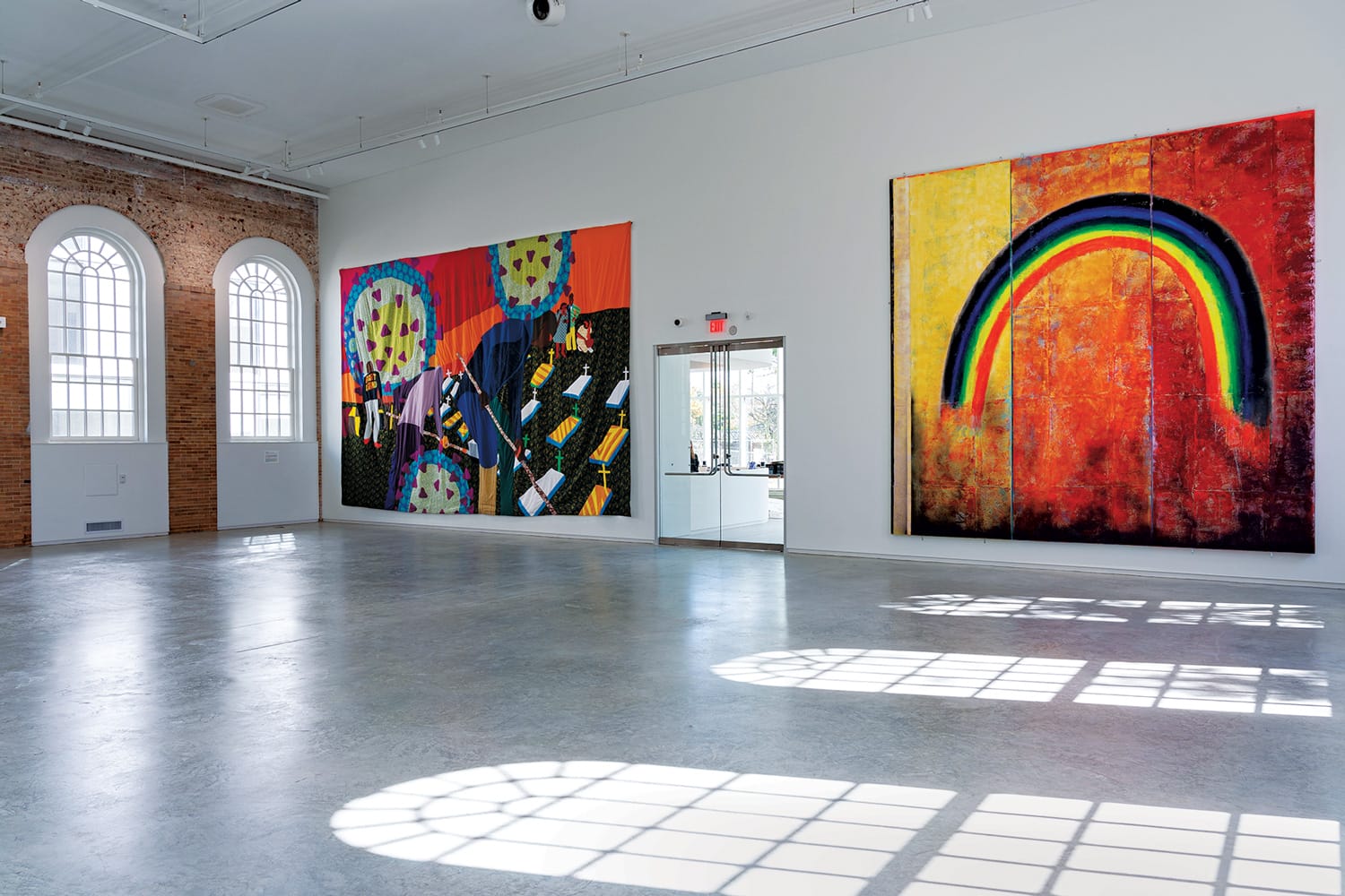 Spacious art gallery with large colorful abstract paintings on white walls and arched windows casting patterned shadows on the floor.