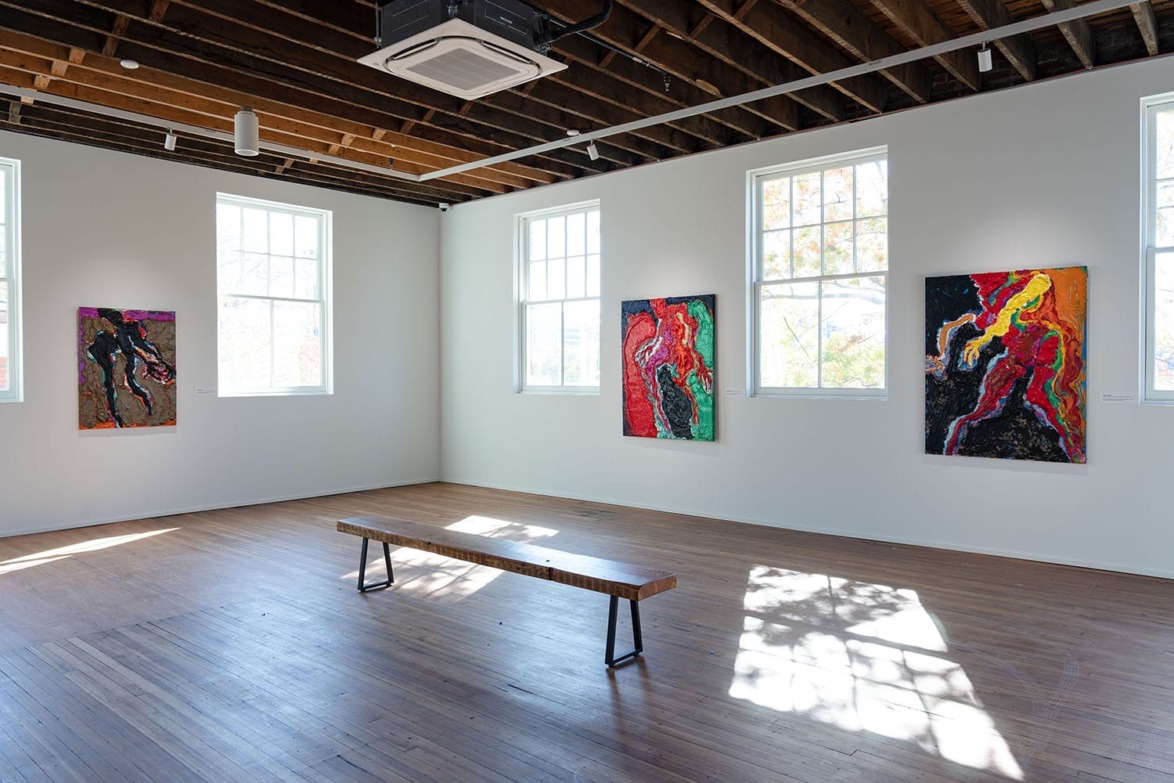Art gallery with abstract paintings on white walls, wooden bench in center, and sunlight streaming through large windows.