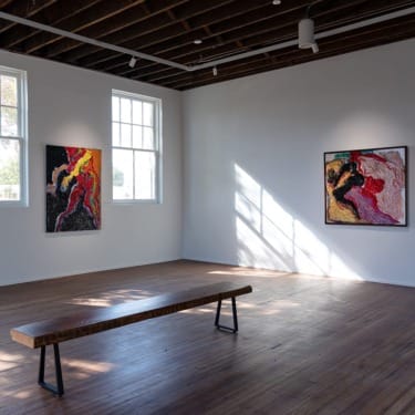 Art gallery interior with two abstract paintings on white walls, wooden bench in the center, and sunlight streaming through windows.