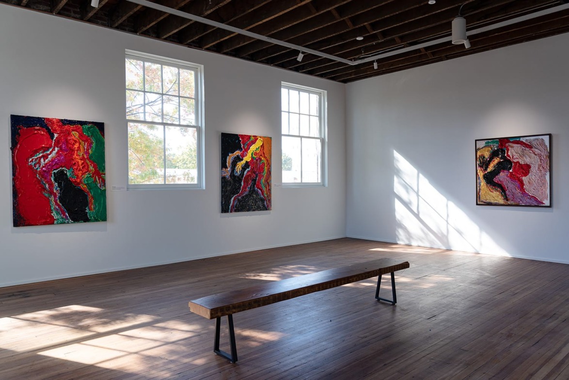 Art gallery interior with colorful abstract paintings on white walls, wooden bench on hardwood floor, and sunlight through windows.