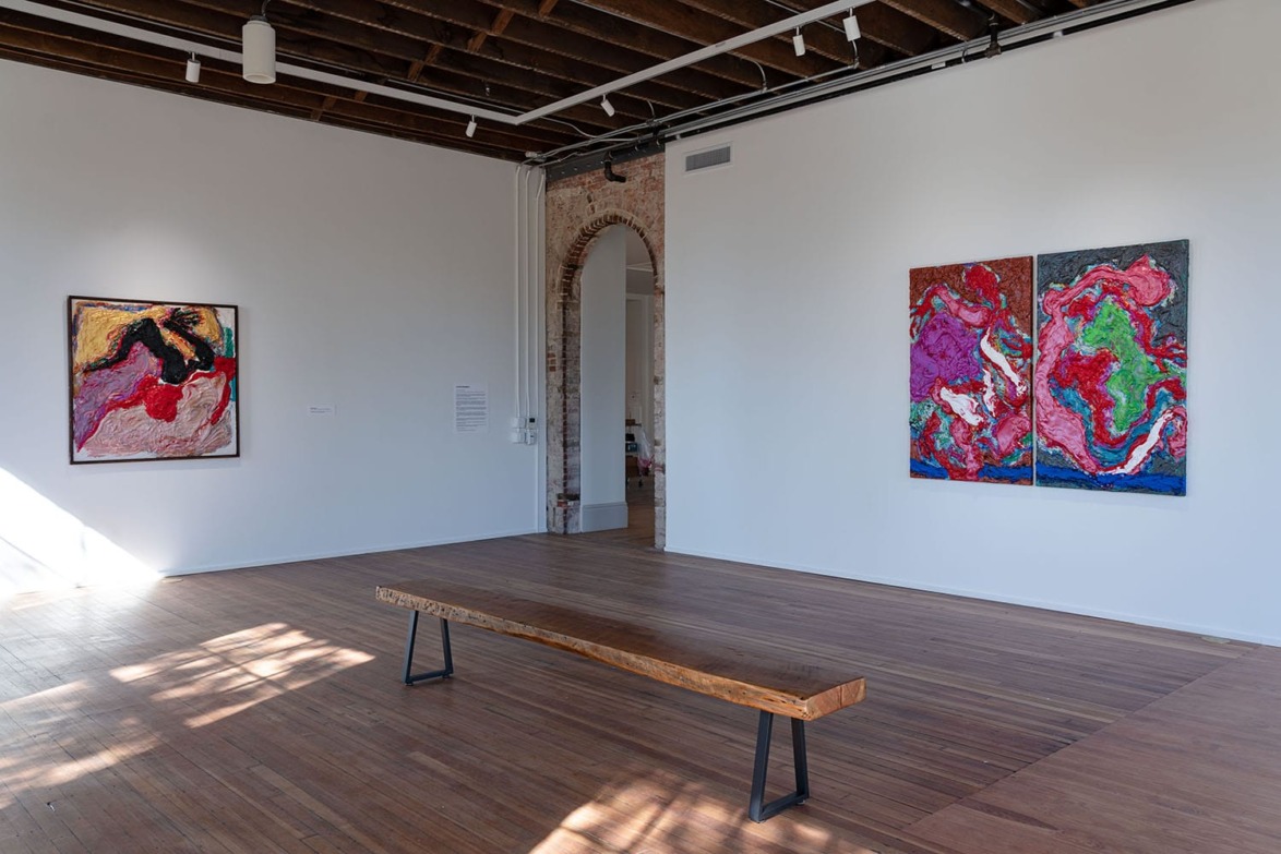 Art gallery interior with abstract paintings on white walls and a wooden bench on the floor.
