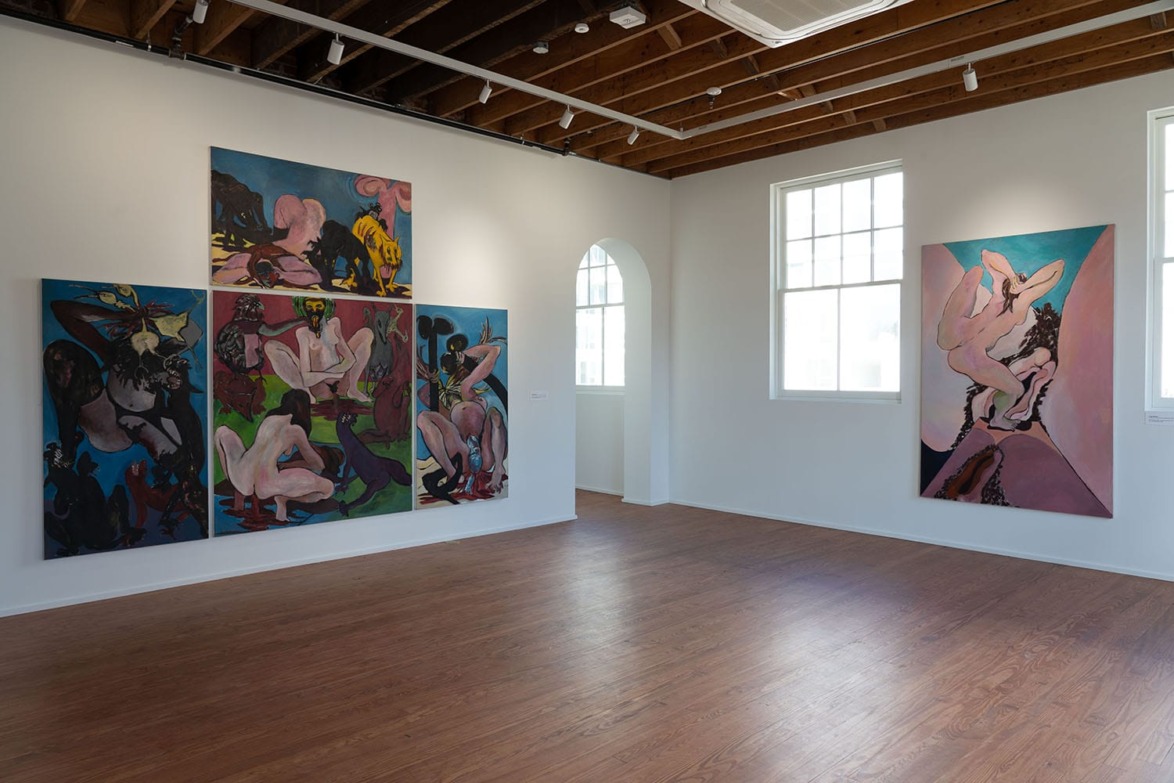 Art gallery with several abstract paintings on walls, wooden floor, and large windows letting in natural light.