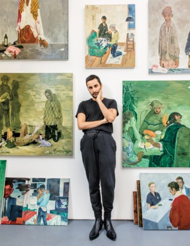 Artist standing in front of a gallery wall displaying various paintings and artworks in a studio setting.