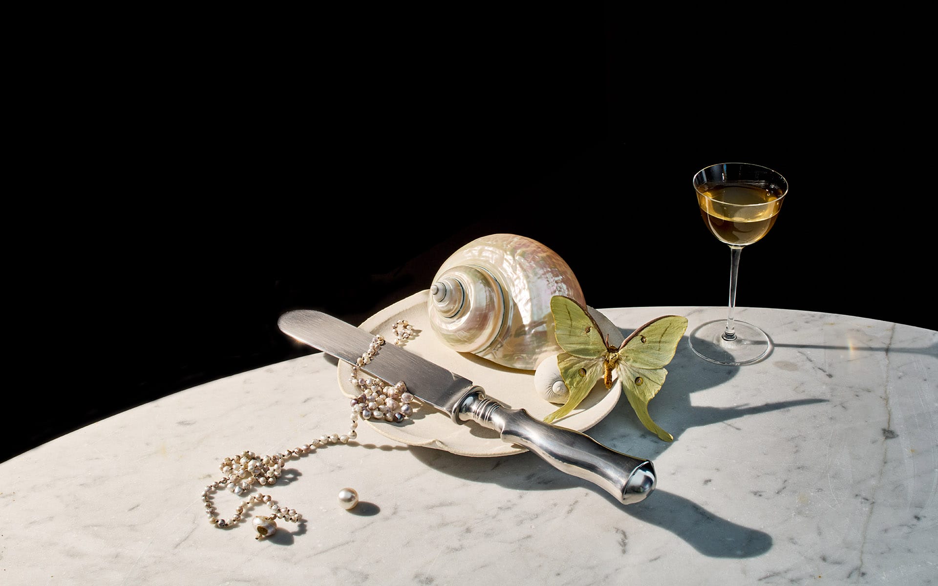 Marble table with a seashell, pearled necklace, butterfly, knife, and glass of white wine against a dark background.
