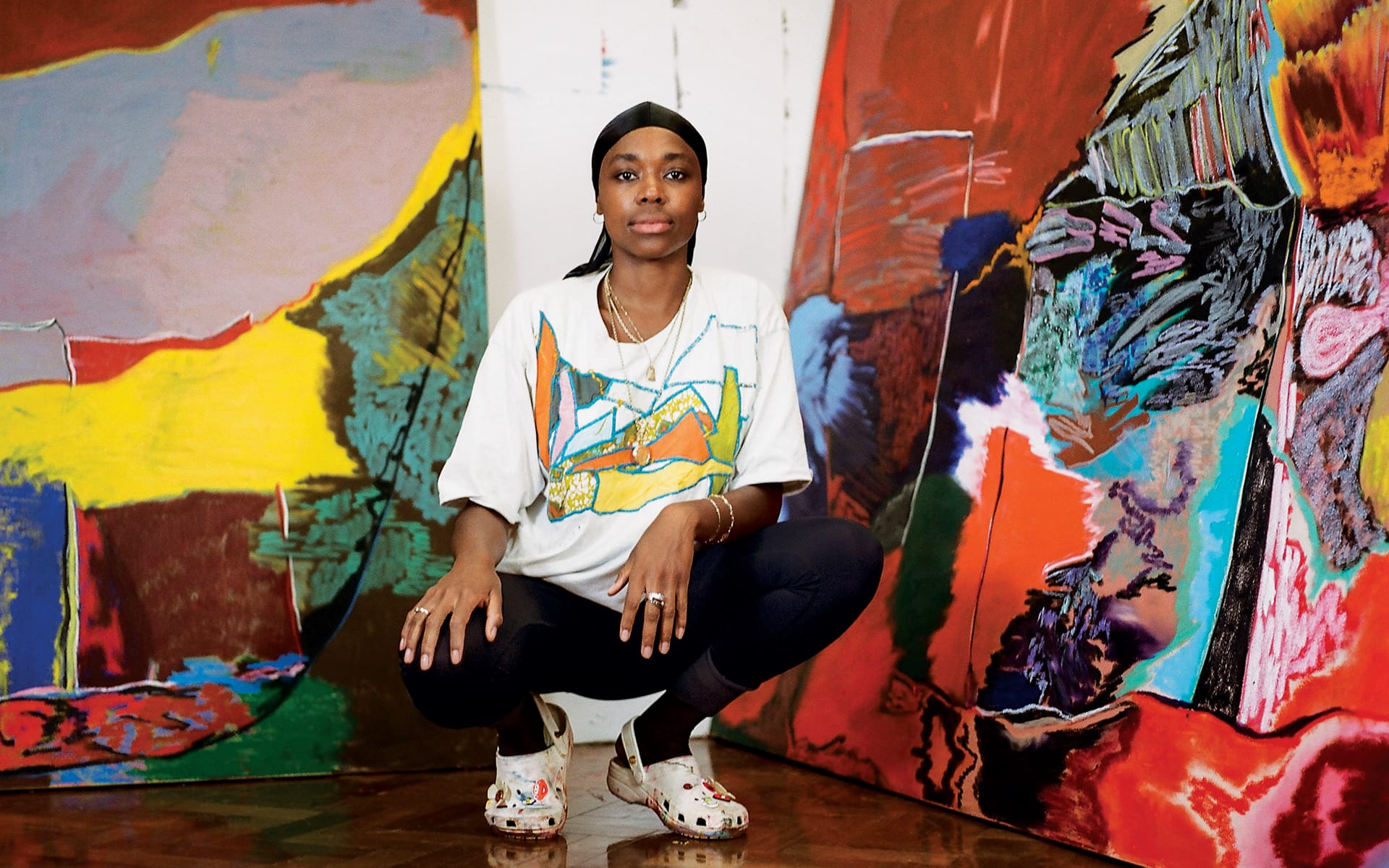 Artist squatting in front of colorful abstract paintings, wearing a graphic t-shirt, black leggings, and white shoes.