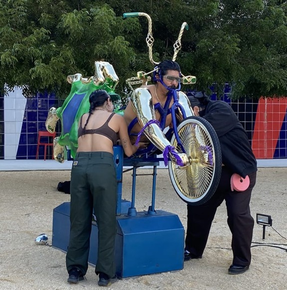 Two people standing with a person wearing a metallic sculpture, resembling a bicycle, against a backdrop of trees and colorful panels.