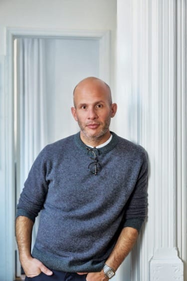 Man in gray sweater leaning against a white wall with glasses hanging from the collar, in a softly lit room.
