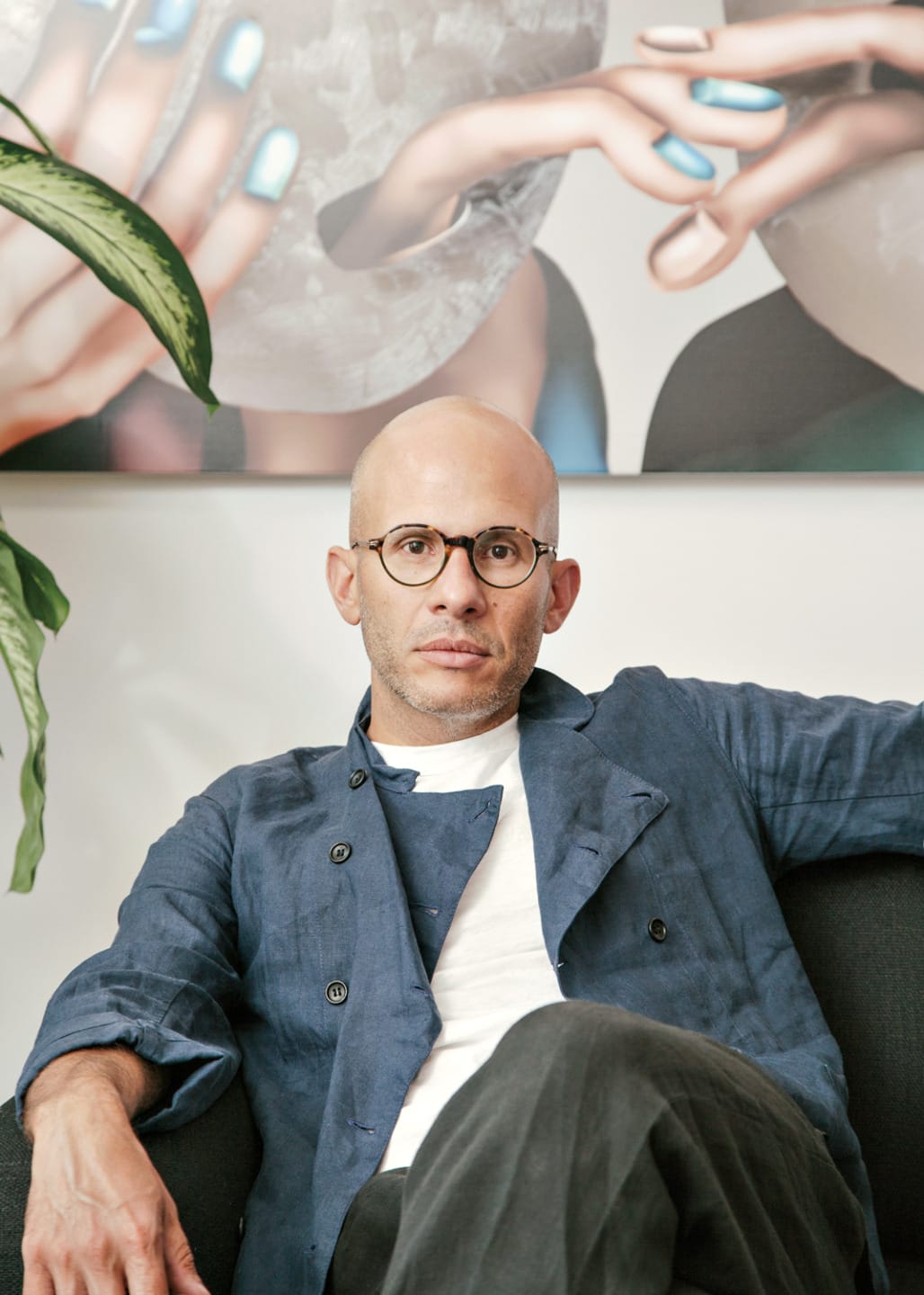 A person with glasses sitting on a chair, wearing a blue jacket with a large artwork behind and a plant beside them.