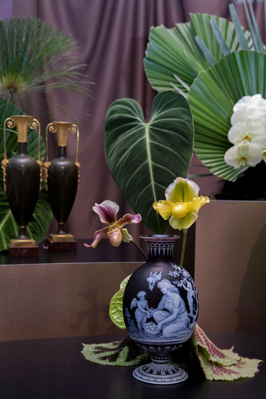 Decorative ceramic vase with a mythological design, surrounded by tropical flowers and large green leaves.
