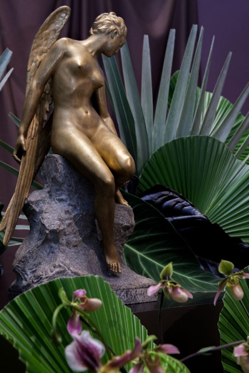 Golden angel statue on a rock surrounded by large green leaves and pink flowers.
