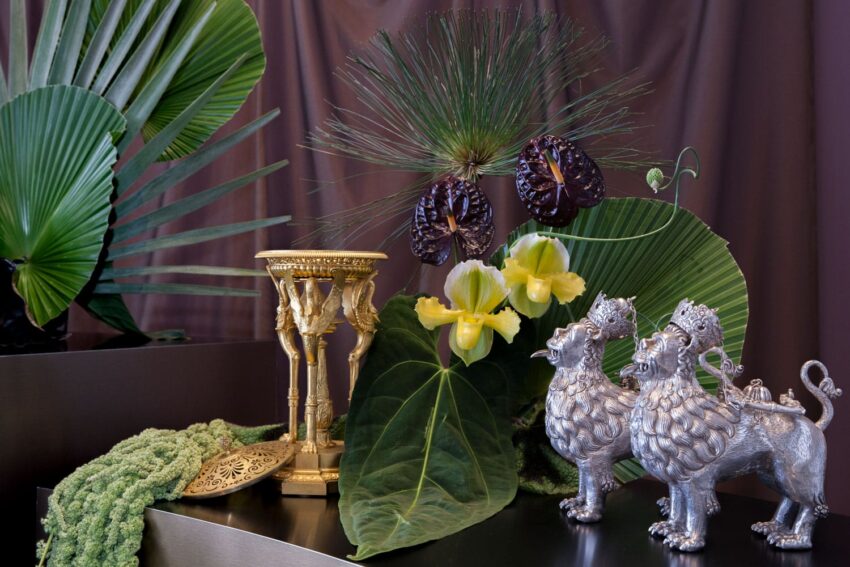 Decorative still life with green leaves, exotic flowers, a golden candlestick, and two metallic lion figurines.