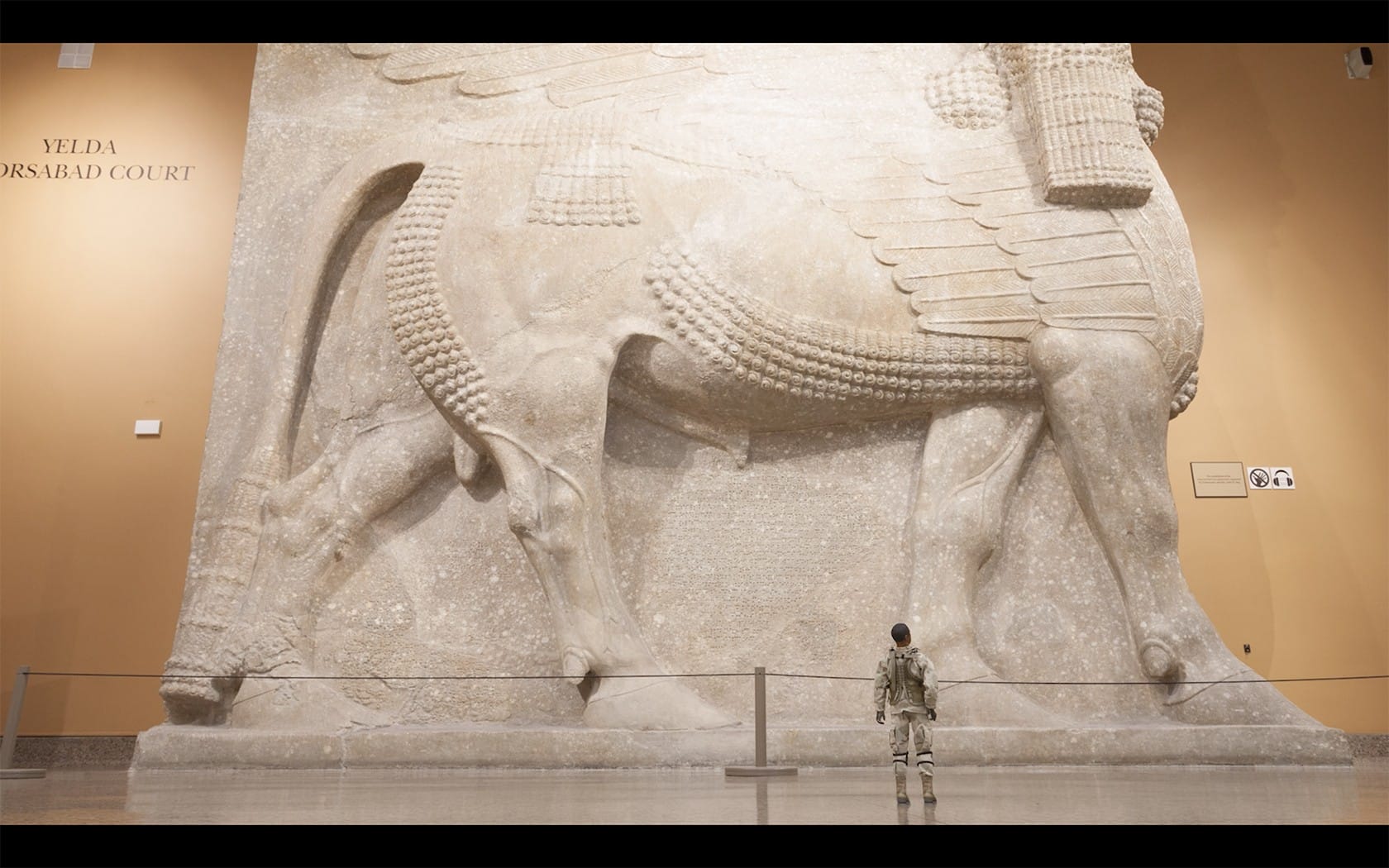 Person standing in front of a large ancient sculpture of a winged bull with a human head in a museum setting.