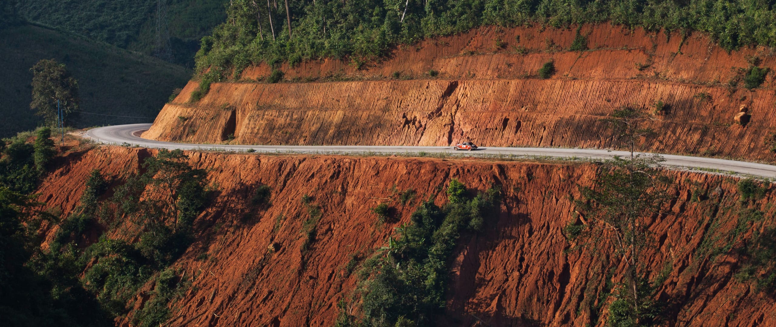 A red car travels on a winding road along a steep, red-earth cliff with lush greenery above and below.