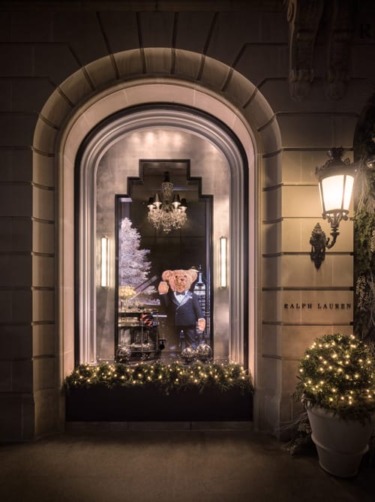 Storefront window decorated for the holidays with a teddy bear in a suit and illuminated garlands around the frame