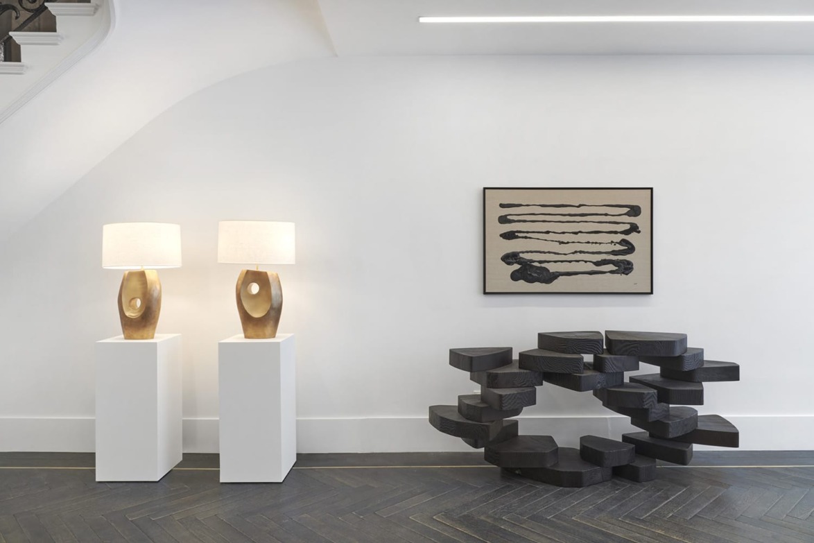 Modern art display with two wooden lamps on pedestals, abstract painting on wall, and unique black sculptural piece.